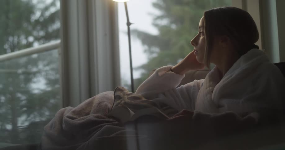 A woman wearing a robe reads a book and looks out window