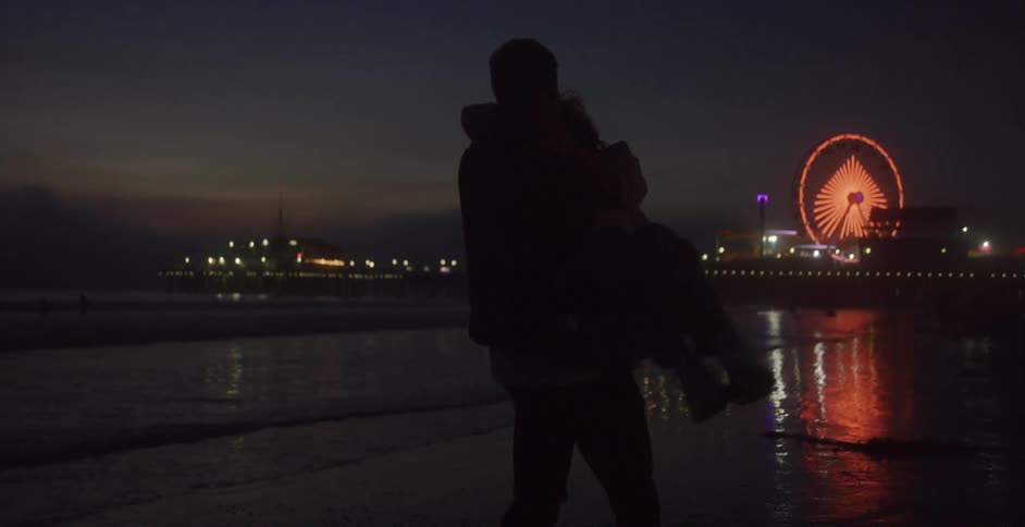 A man walking on the shore carries a woman in his arm and spins her around with the city lights in the distance