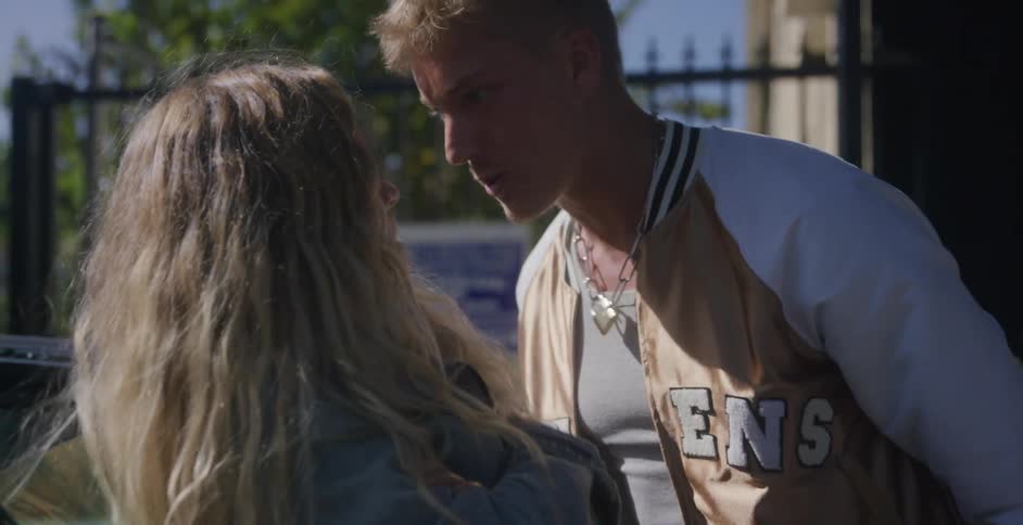 A teenage boy walks around a car and yells at a girl leaning on it and grabs her face and continues yelling at her