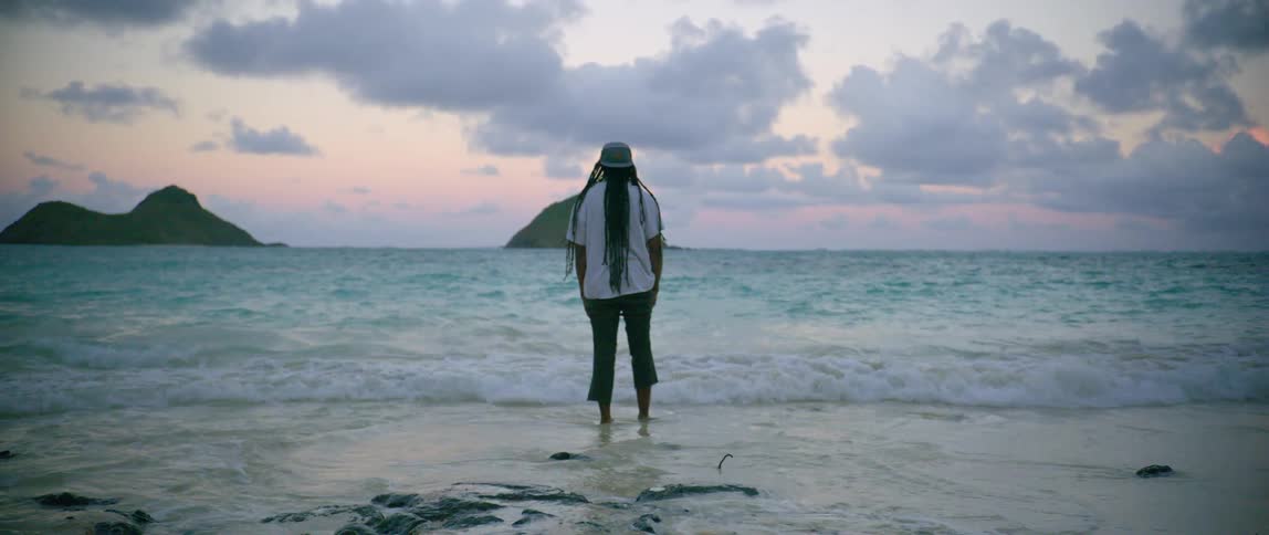 A man with a hat standing on a beach with the sky and clouds in the background
