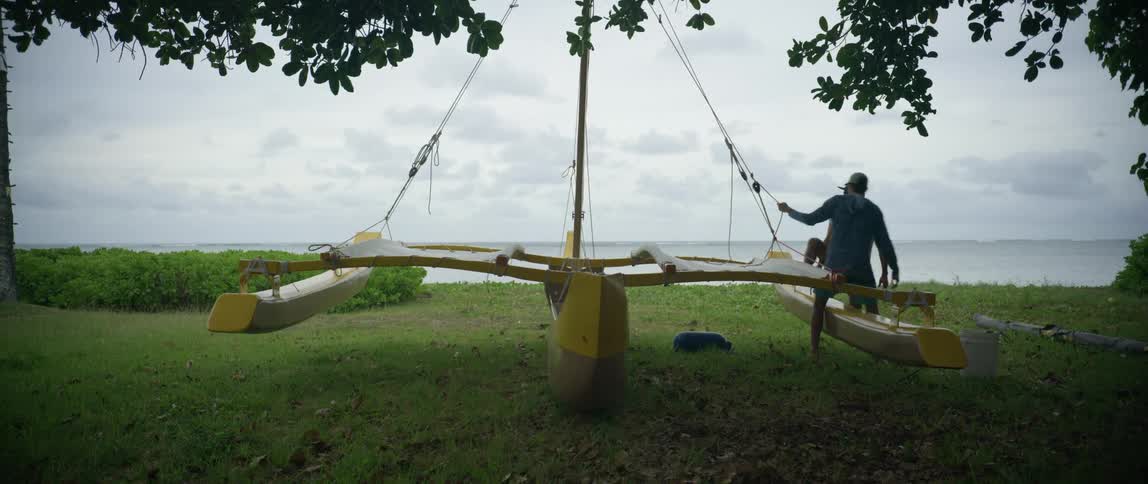 Two men work on the rope lines of a catamaran on the grass shore