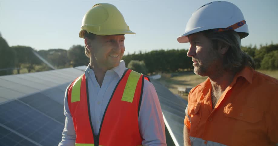 Two men with hard hats on stand at a work site and talk