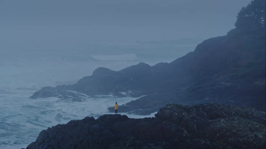 Aerial shot pulls away to reveal woman standing on rocks as ocean waves crash around her