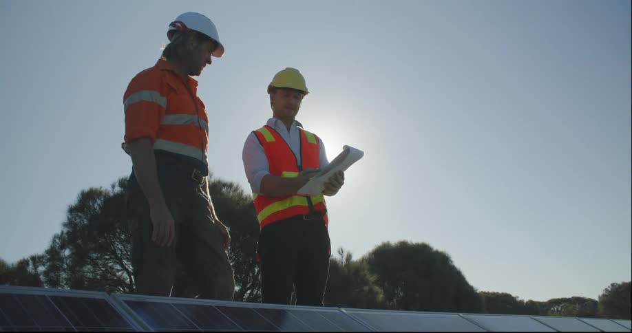 A man with a clipboard talks to another man wearing safety gear standing on a roof with solar panels at daytime
