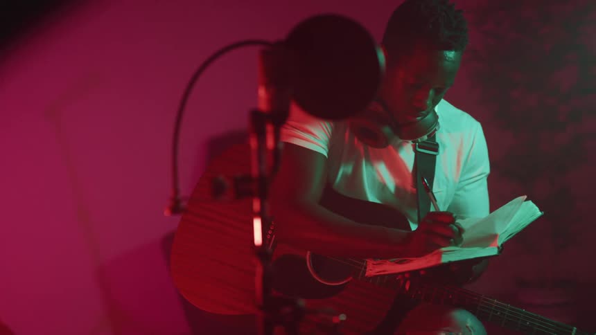 A black man sitting with a guitar writing in a journal with a microphone in front of him
