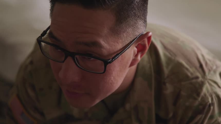 A soldier sitting in a military uniform and glasses looks up from the floor
