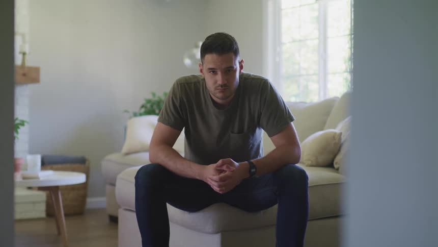 A man sitting in a room on a couch with a wall and window in the background