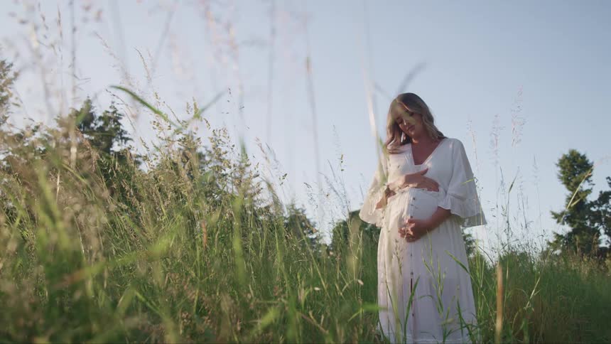 A pregnant woman wearing a white dress is holding her stomach as she stands outdoors in a field