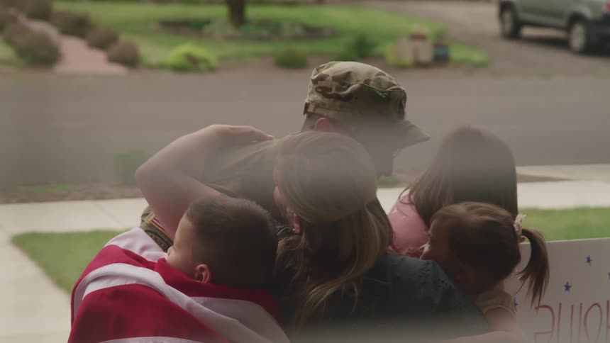 A woman and children hugging a man with a military uniform and glasses with a street and a vehicle in the background