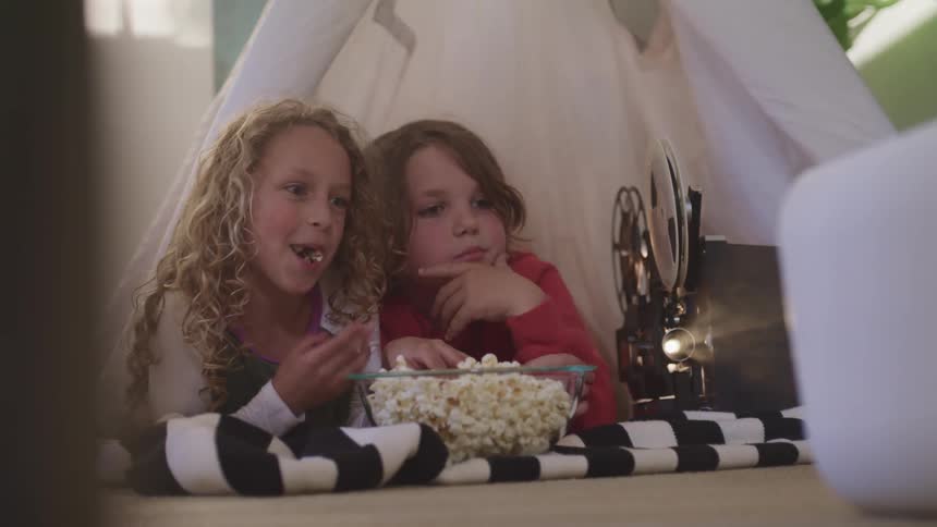 A boy and girl lying side by side in a play tent in a house eat popcorn while watching a film being displayed by a projector