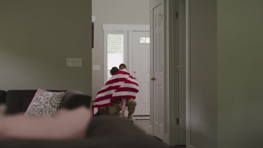 A soldier returns home and is greeted with a hug from his daughter wearing an american flag