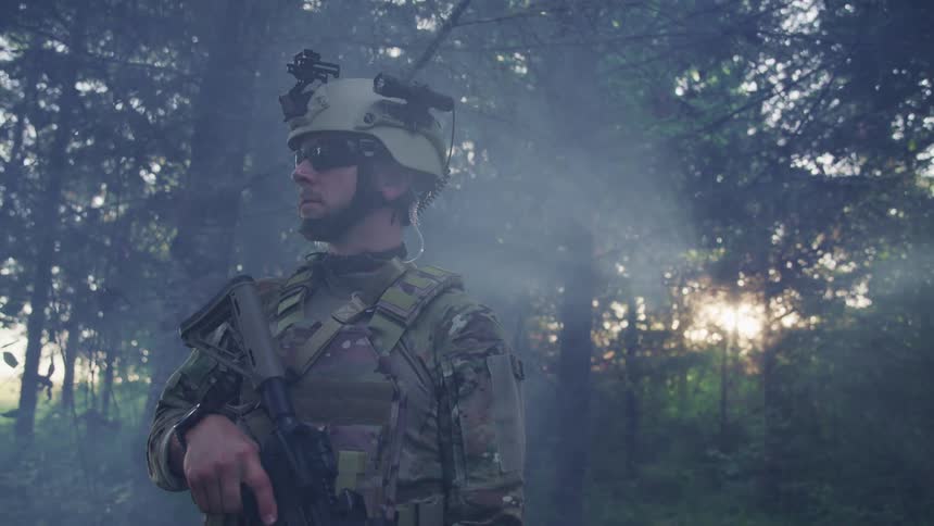 A soldier standing in a forest holds a gun while the sun beams through the trees behind him