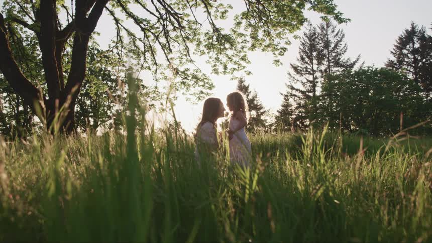 Daugther kisses mother forehead while standing on the tall grass of a field at sunset