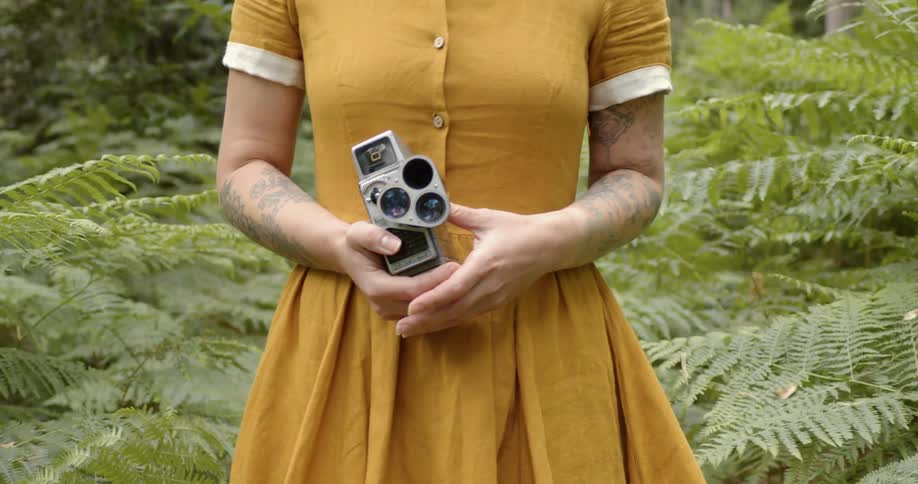 A woman walks backward through a forest with a film camera