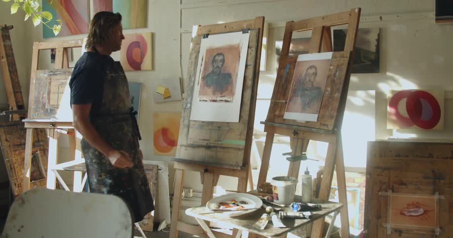 A tall man pauses and looks at his painting before leaning over to get more paint on his brush