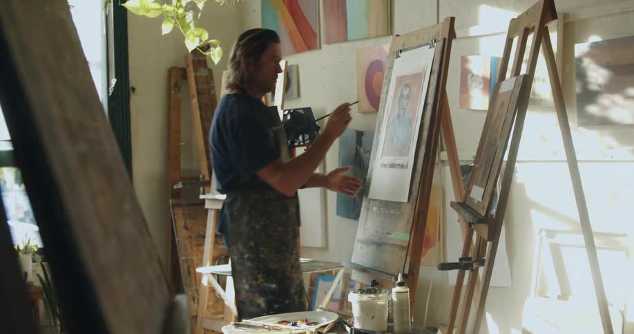 Man stands back and looks at a picture next to an easel with a painting he is working on then moves closer and strokes a paintbrush