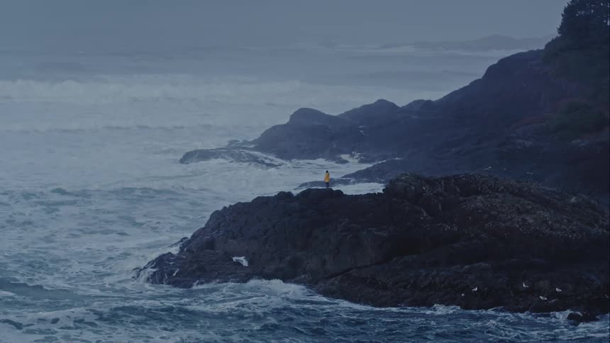A woman standing on an exposed rock as stormy ocean waves crash around her