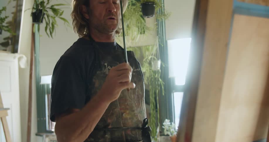 A caucasian male wearing an apron painting on a canvas in a studio with lots of plants and windows