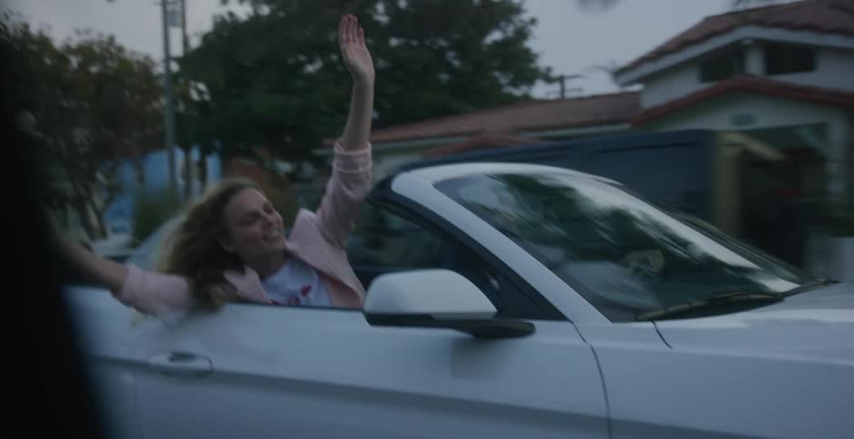 A woman and man driving through a neighborhood in a convertible with their arms outstretched feeling the wind