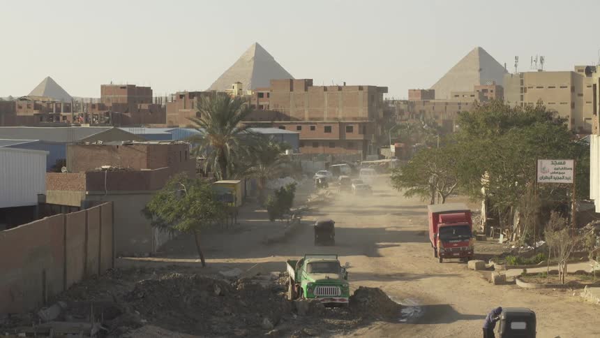 Cars and trucks travel on a main street dirt road with pyramids standing tall behind the buildings