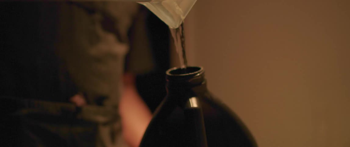 A person pouring a liquid from a plastic jar into a dark bottle