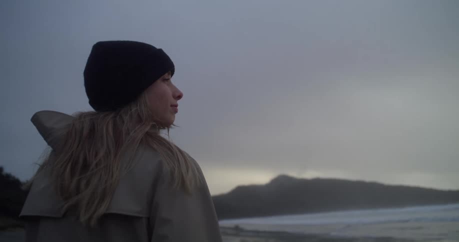 Young long haired blonde woman wearing jacket and beanie looking at the windy ocean at sunset