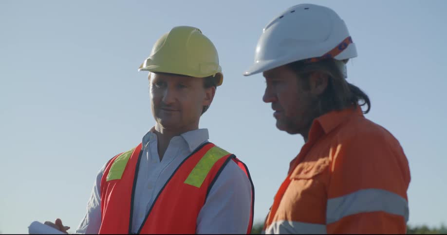Two men wearing construction gear standing outside communicating with one another