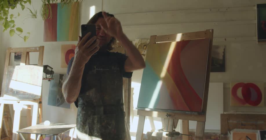Man wearing an apron on a video call on his cellphone showing his painting