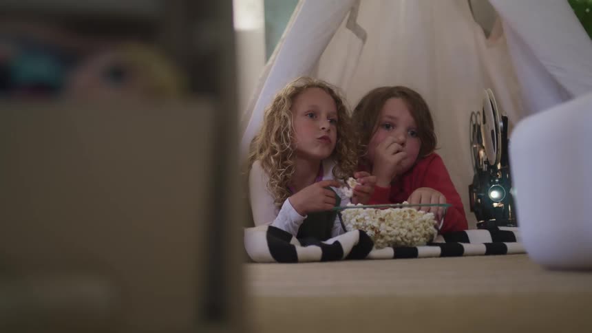 Boy and girl eating popcorn next to a film projector inside a camping tent