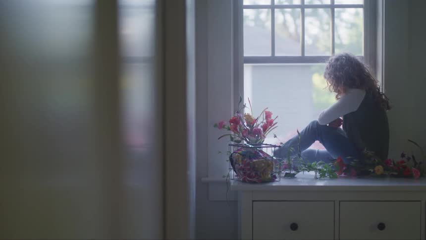 A girl gazes out of a window while sitting atop a dresser in a house during the day