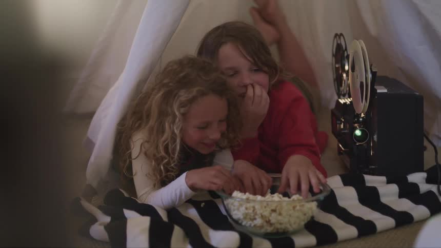 A boy and girl lying in a play tent in a house eat popcorn while watching a film being displayed by a projector together