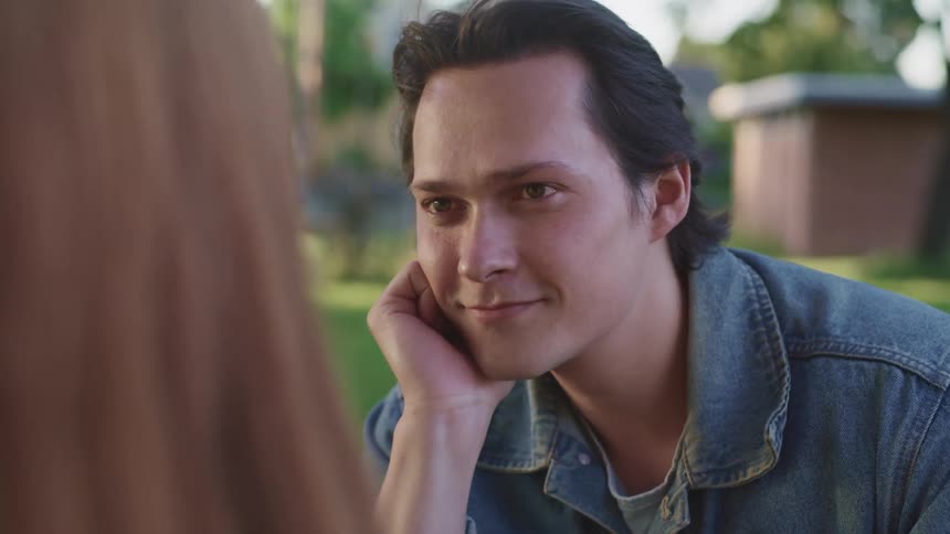 A man sits with his head resting on his palm as he smiles at his girlfriend while sitting in a park