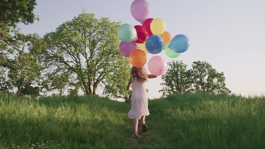 Young girl in a dress running through a grass field with colorfull balloons at sunset