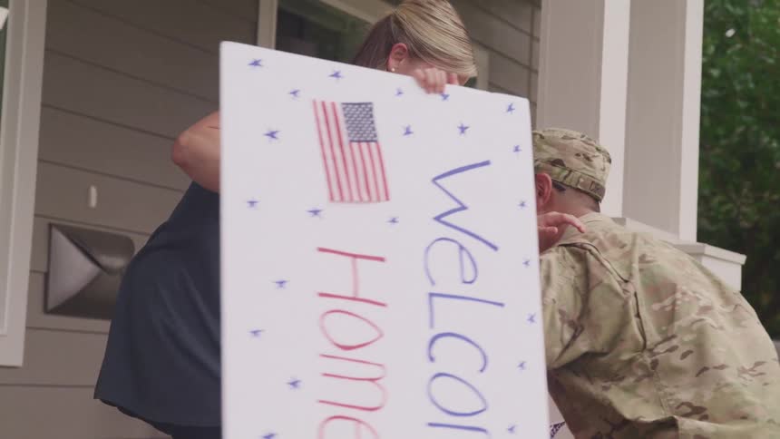 Women and children standing on a porch in front of a house hugging a soldier returning home