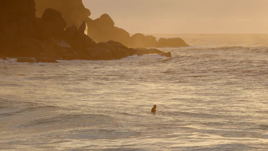 Man swimming in ocean waves next to large rocks and a mountain at sunset