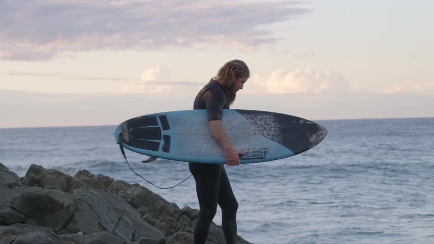 A bearded man wearing a wet suit is carrying a surfboard towards the ocean