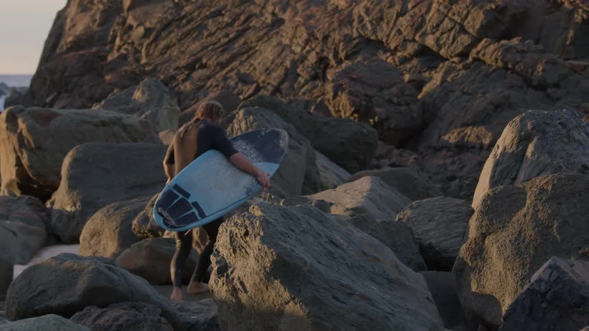Man holding a surfboard climbs up rock formations on a beach in the evening