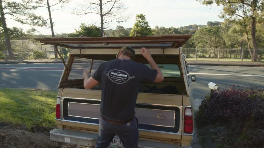 A bearded man places a bag and a wet suit in the back of his truck