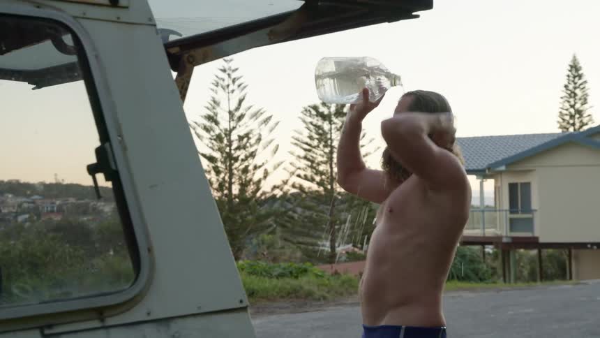 White man picking a water bottle and pouring the water in his hair and body next to a car