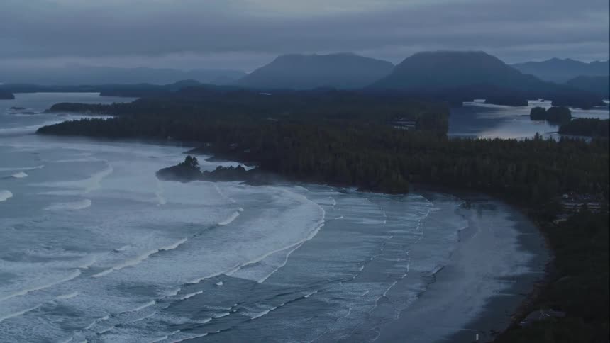 Waves roll in over beach with a rocky coastline and mountains in the background and a cloudy sky