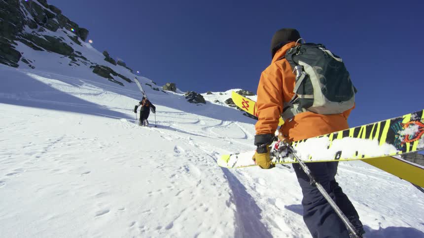 Skier carries skis up a snowy mountain while another skier skis up the mountain ahead of him