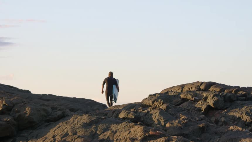 A man with a long beard in a wet suit walking along some rocky hills with a surfboard