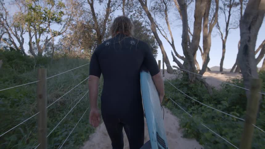 Man holding a light blue surfboard walks up a narrow trail towards the beach at daytime