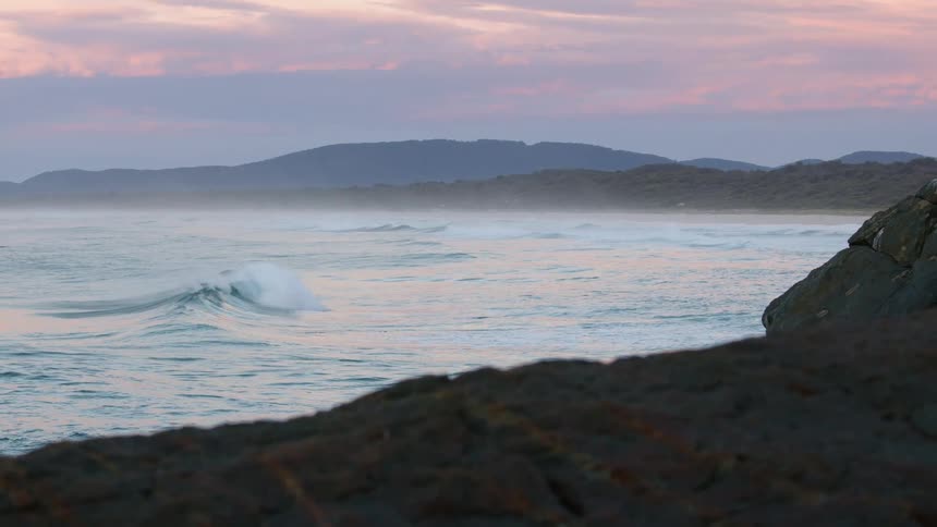 A wave slowly rolls towards a coastal shore lined with rocky terrain
