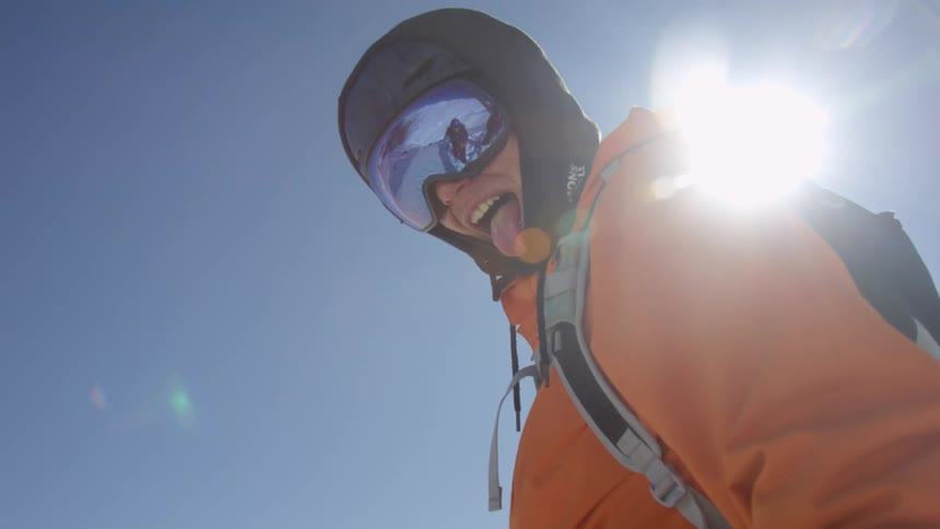 A teenager standing out in the snow with a hat and goggles on smiles and sticks their tongue out