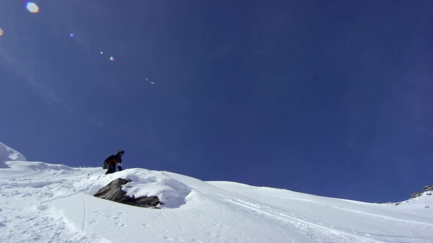 Skier jumps over a snow covered rock on the slope of a mountain on a clear day with blue sky