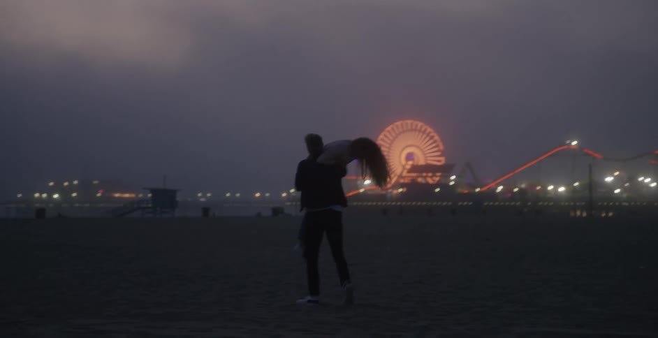 A man runs toward his girlfriend on the beach and picks her up hear the boardwalk