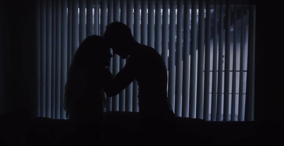 Man and woman embrace in a dark room with blinds on the window