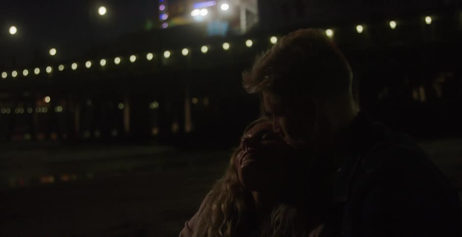 Man holds blonde woman and kisses her lovingly next to sea at night