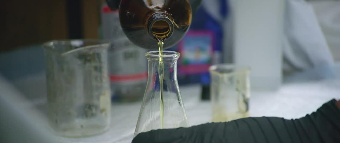 A person wearing gloves pouring a yellowish liquid into a beaker